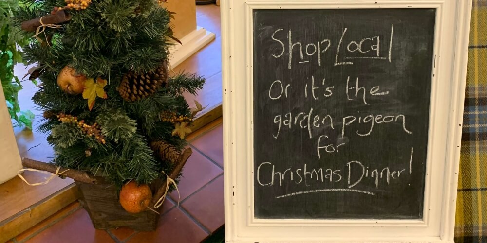 A hand-chalked Shop Local sign on a white-framed blackboard inside the shop, next to a Christmas planter and a tartan throw